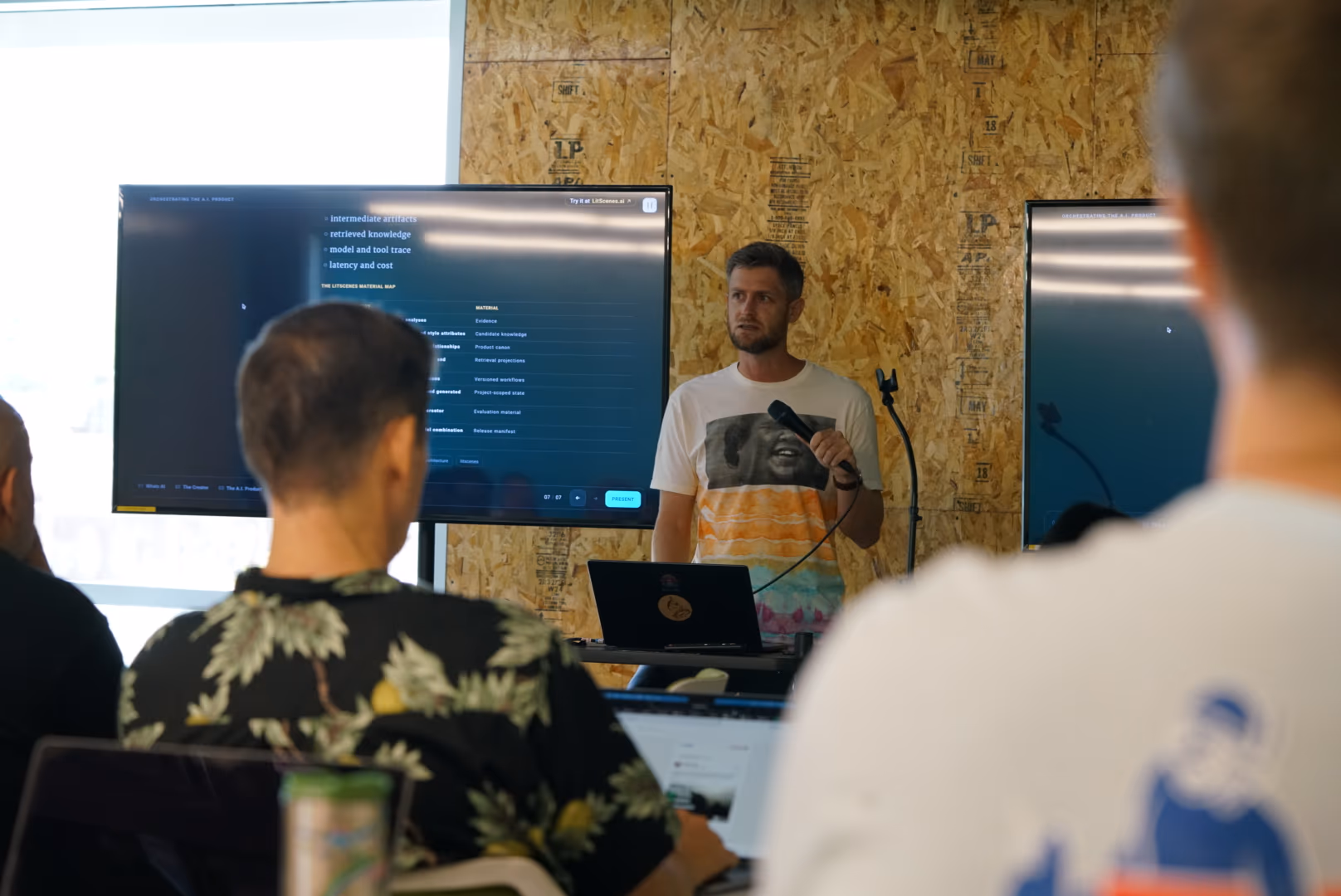 A speaker presents at a laptop between two displays, with seated participants in the foreground.