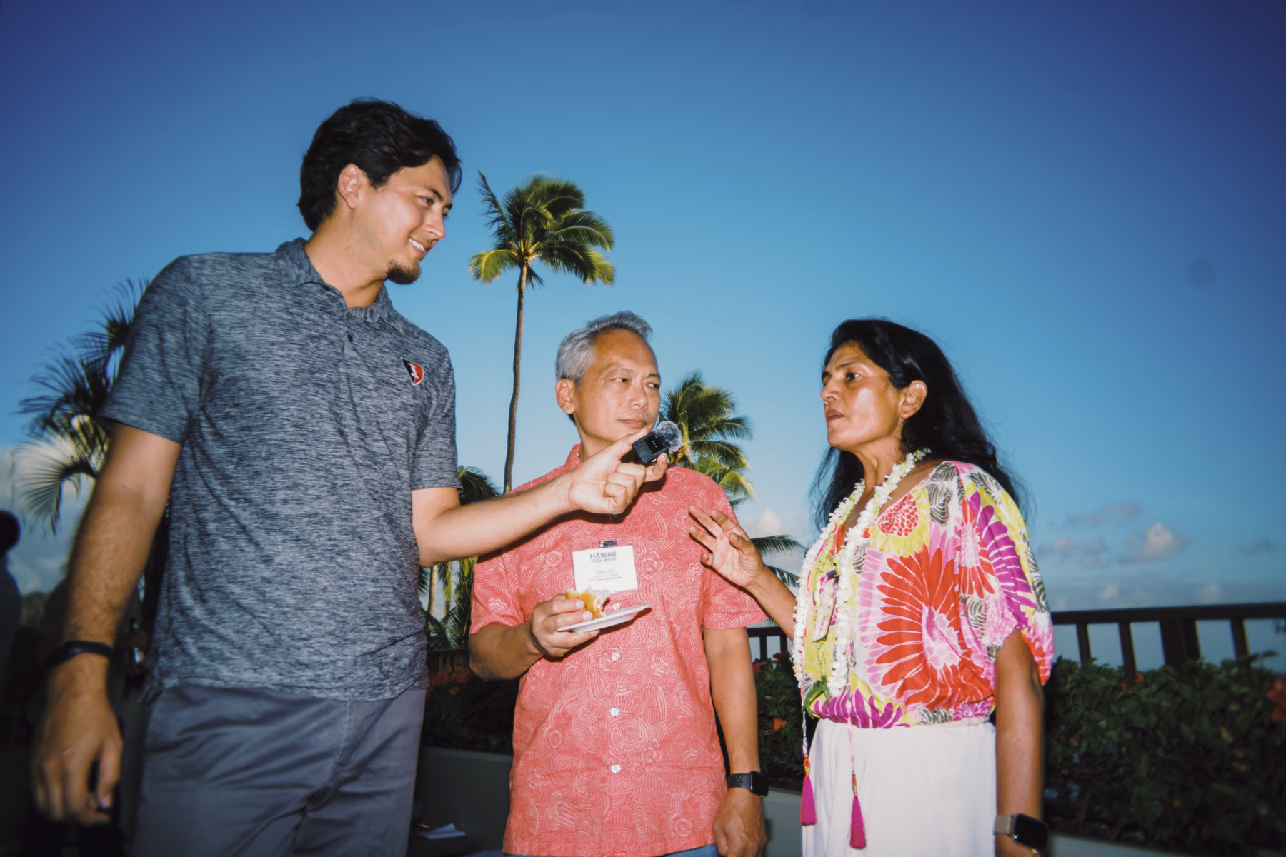 Three participants talk on an outdoor terrace during the Executive Forum, with a microphone, palm trees, and the ocean behind them.