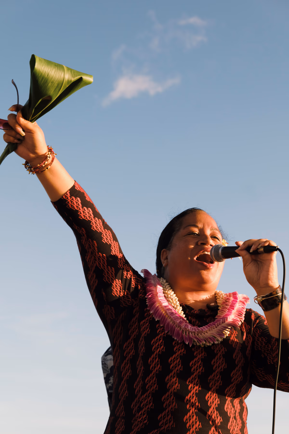 A kumu holds a microphone and raises green leaves during a chant aboard the sunset sail.