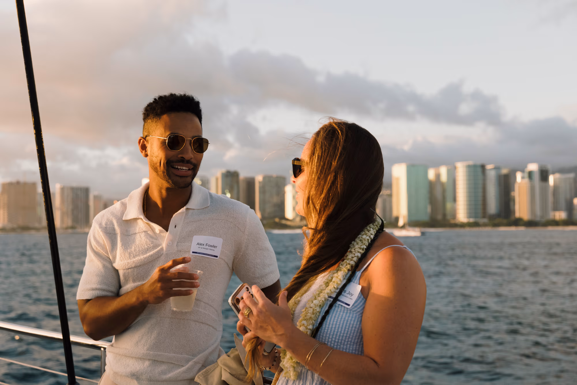 Two people converse on a boat with the ocean and Honolulu skyline behind them.