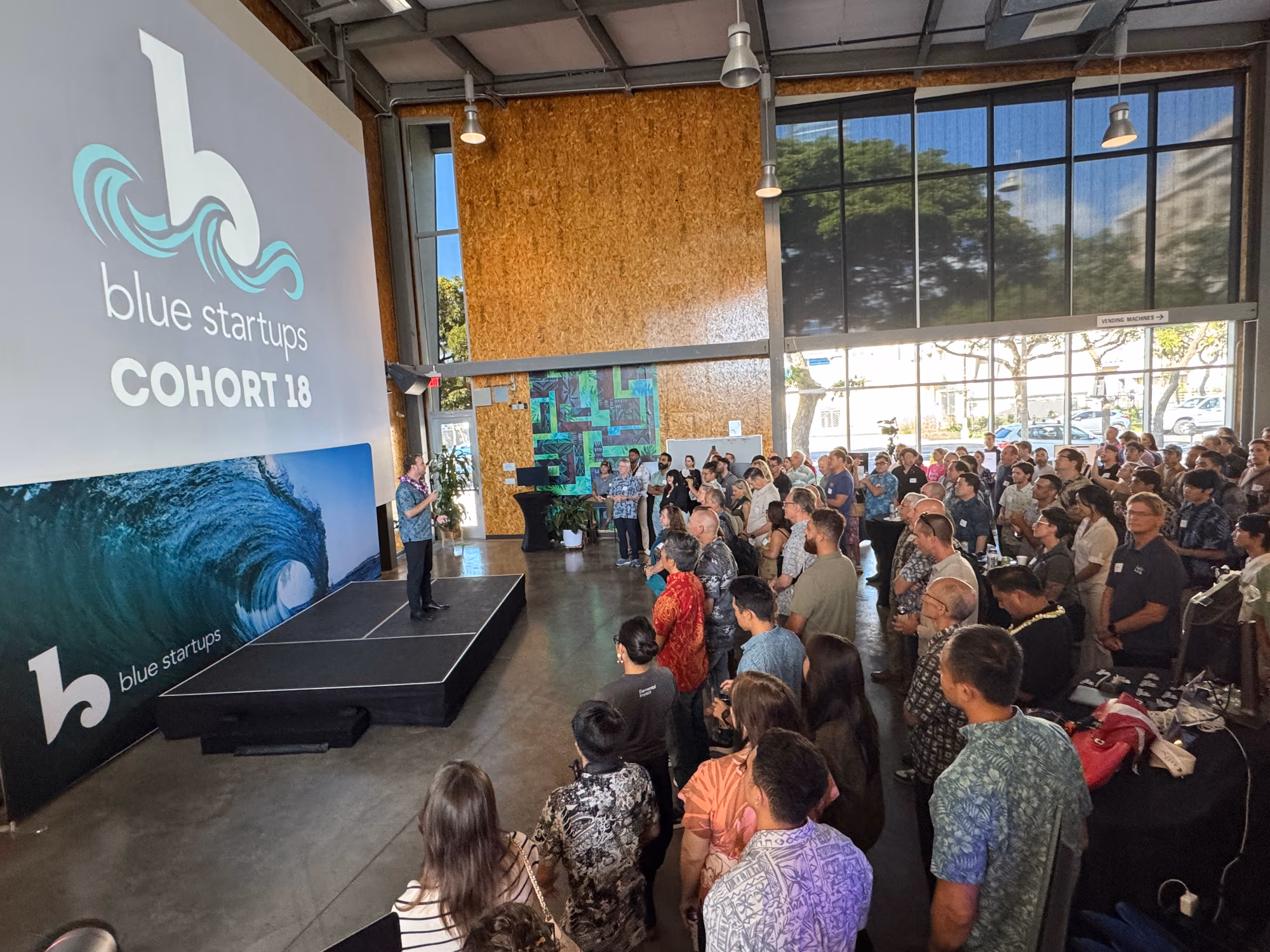 A speaker addresses a full audience beside Blue Startups branding at its 2026 Cohort 18 Demo Day