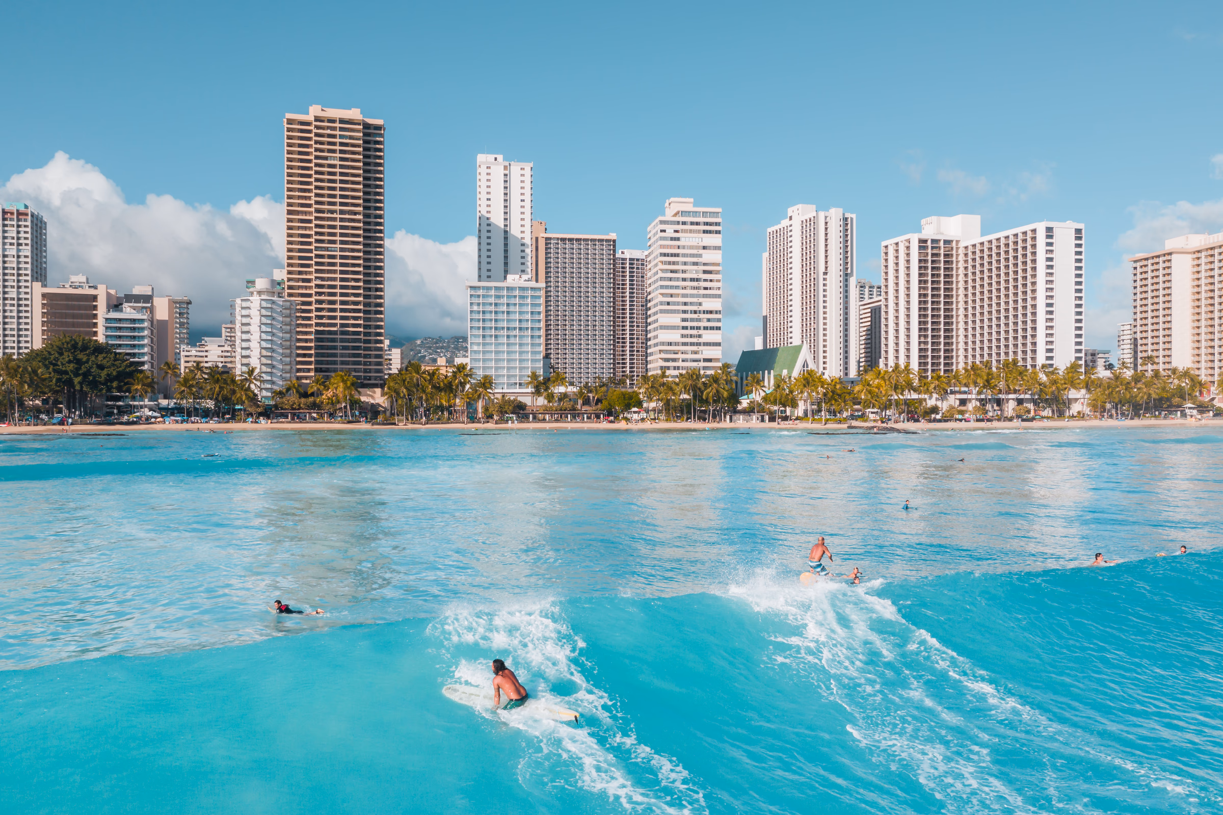 Sunny Waikiki coastline with bright blue ocean, surfers, hotel towers, and palms
